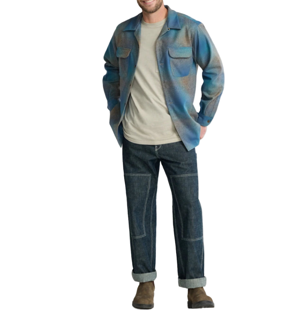 Man wearing a blue and gray shirt, beige t-shirt, and dark jeans on a light background