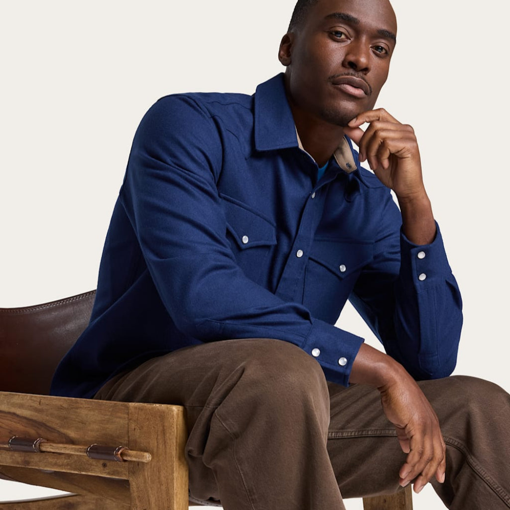 Man sitting on a wooden chair wearing a blue shirt and brown pants.