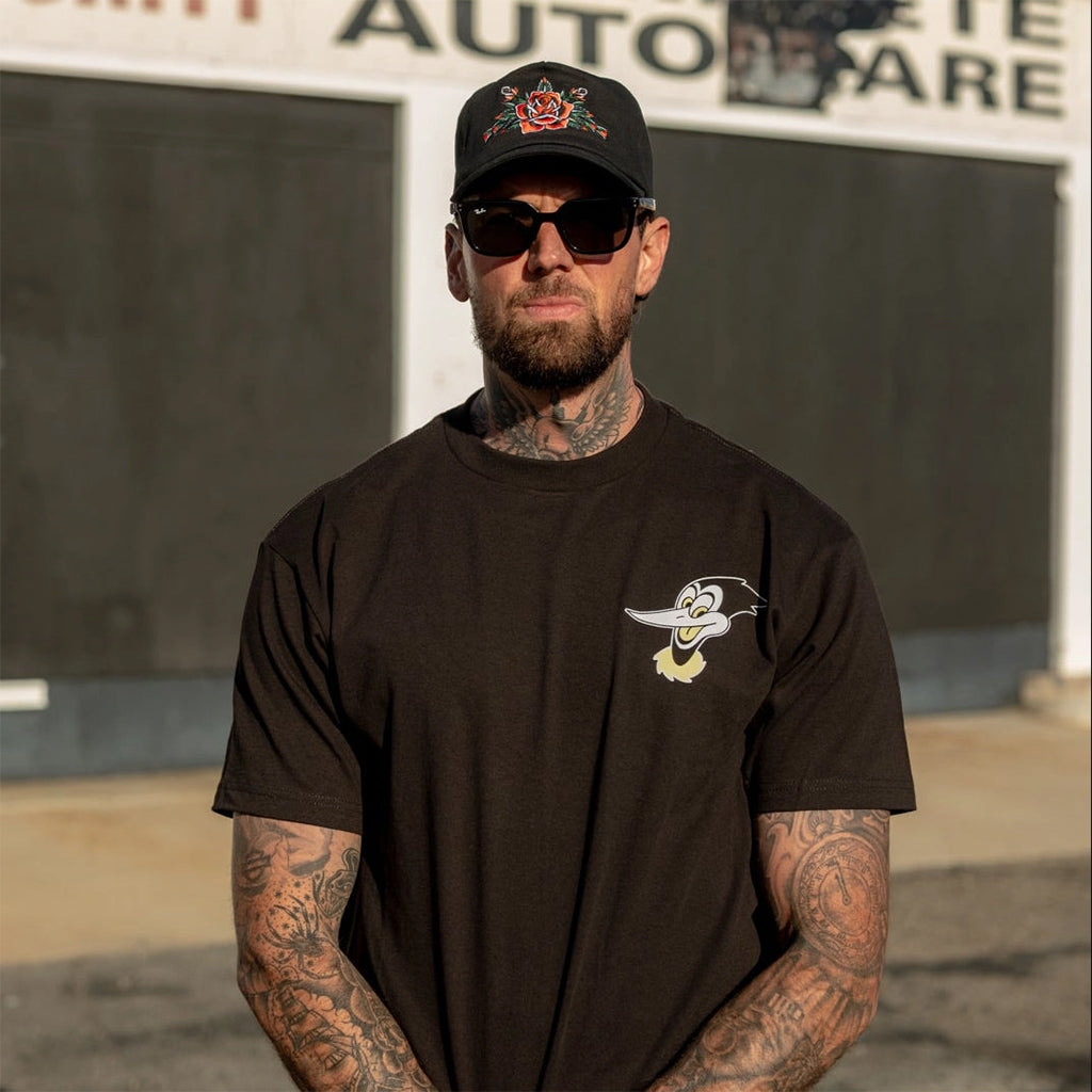 Man wearing a black t-shirt with a logo and a cap, standing in front of a building with text.