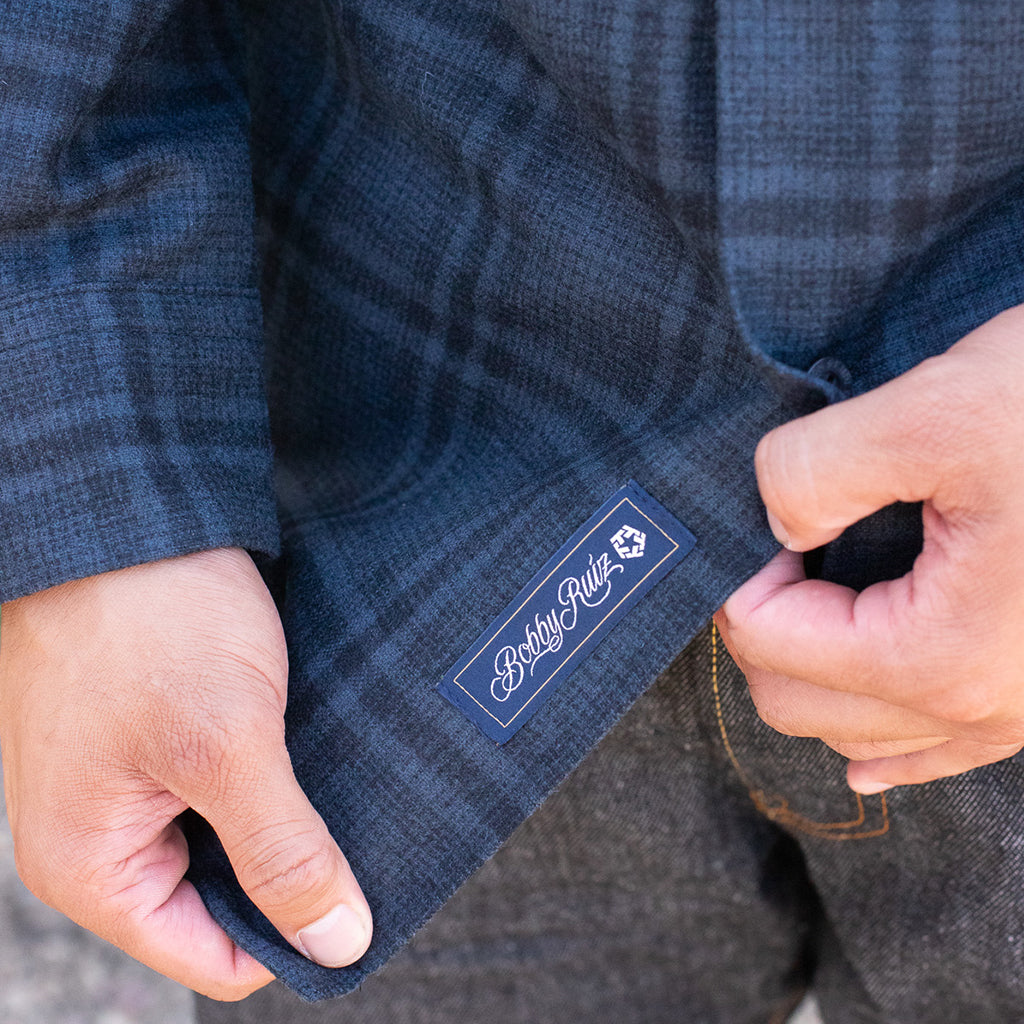 Close-up of a person adjusting a blue plaid shirt with a brand label, wearing brown pants.