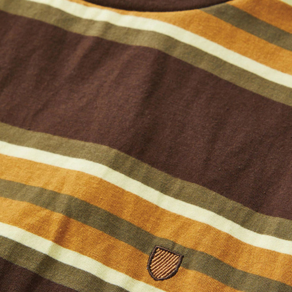 Close-up of a striped fabric garment, likely a sweater or knit top, with horizontal bands in dark brown, mustard yellow, olive green, and cream. A small shield-shaped embroidered patch is visible near the lower center.