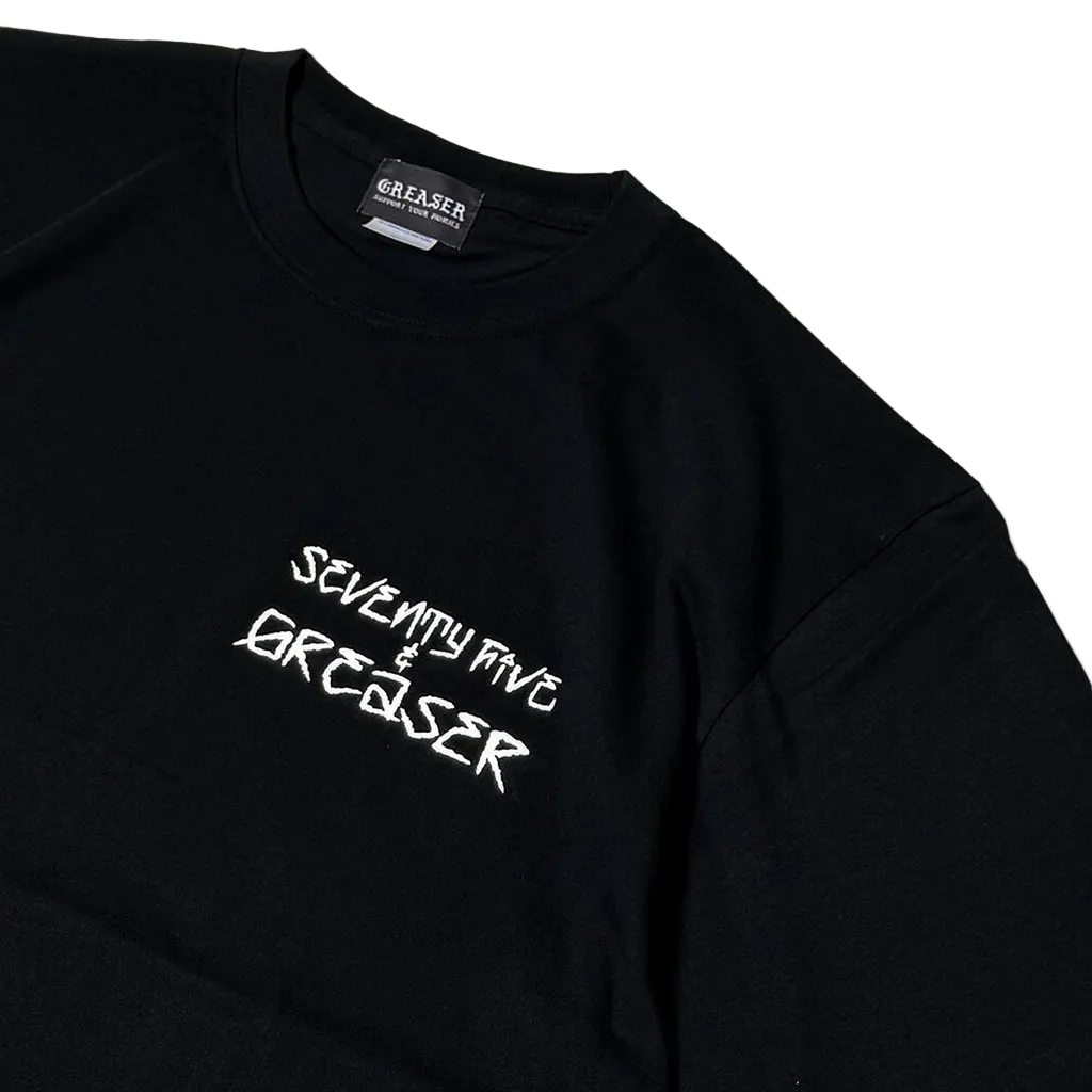 A close-up of a black long-sleeve t-shirt with 'SEVENTY FIVE & GREASER' printed in white stylized text on the chest. The brand label 'GREASER' is visible inside the collar, and the photo is shot on a clean white background.