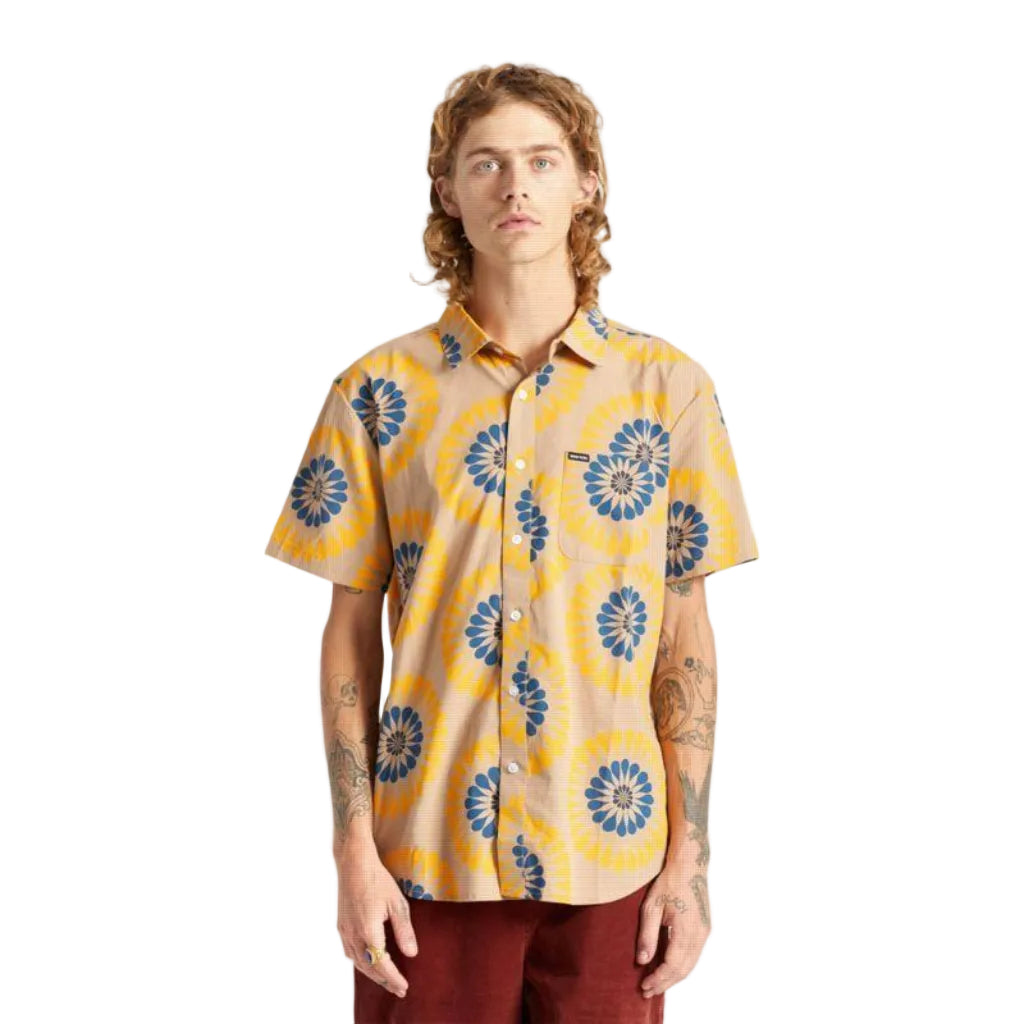 A man wearing a short-sleeve button-up shirt with a beige base and a yellow and blue sunburst pattern, paired with dark red pants, standing against a plain white background.
