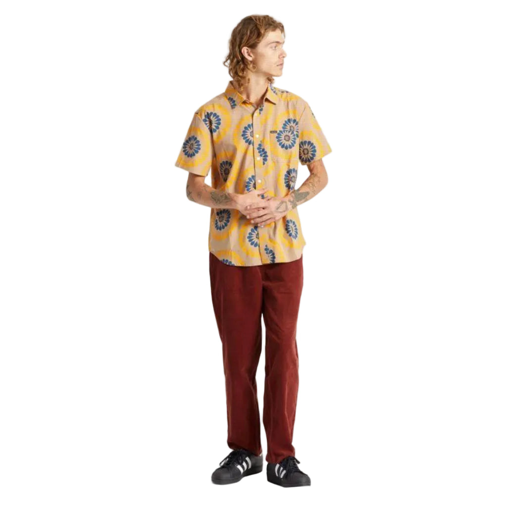 A man wearing a yellow short-sleeve button-up shirt with a blue and yellow floral pattern, paired with rust-red corduroy pants and black sneakers, standing against a plain white studio background.