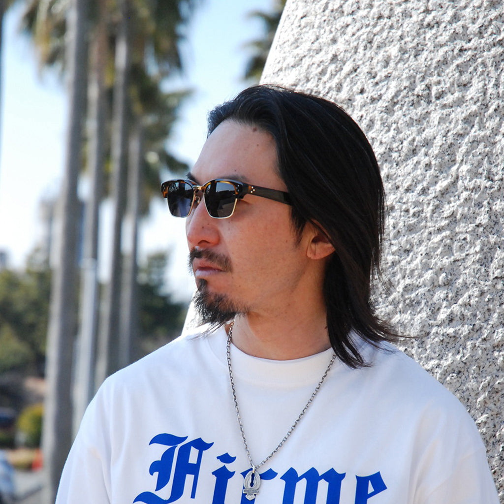 Man wearing sunglasses and a white sweatshirt with blue text, standing against a textured wall.