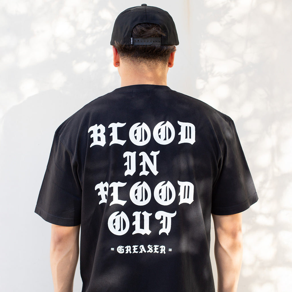 Person wearing a black t-shirt with 'Blood In Blood Out' text on the back, against a light gray background.