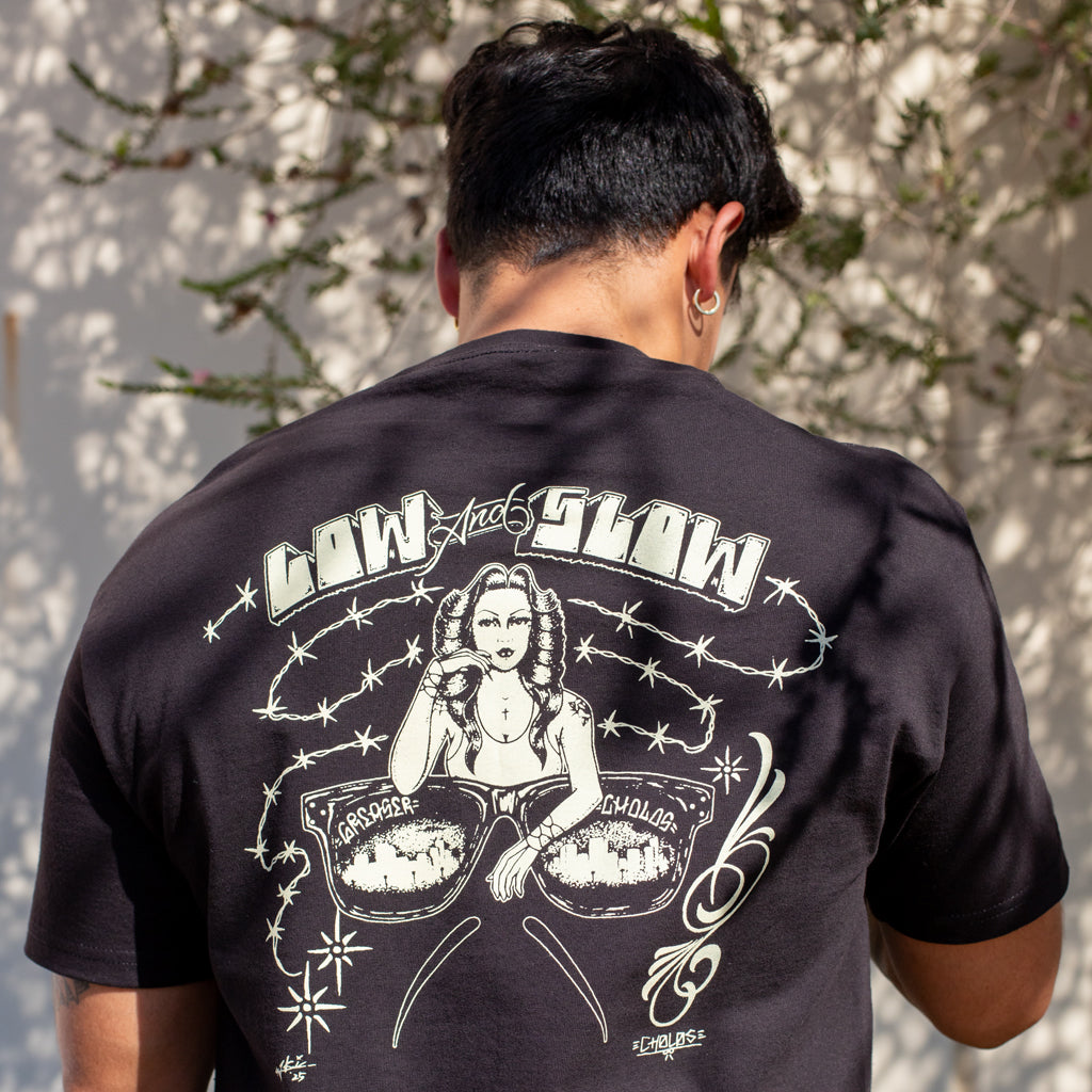 Person wearing a black t-shirt with graphic design and text on the back, standing against a light-colored wall.