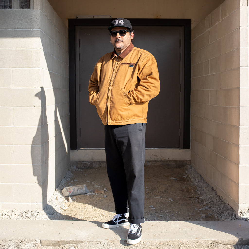 Man wearing a mustard jacket and black cap standing in a sunlit outdoor area.
