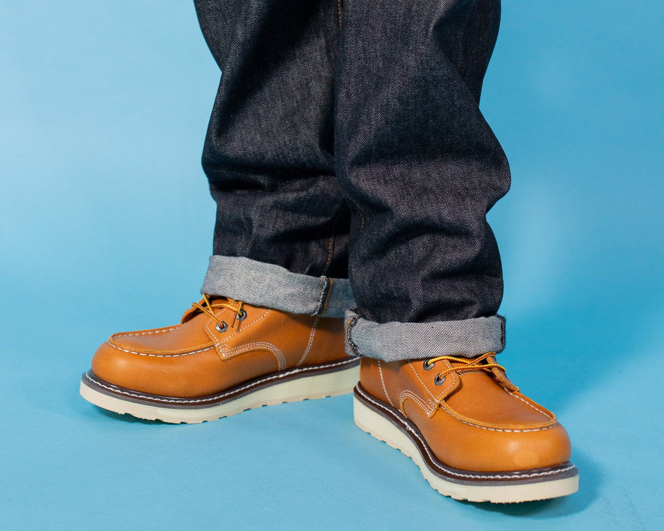 A close-up of a person's legs wearing dark indigo denim jeans with rolled cuffs and tan leather lace-up shoes, set against a solid light blue background.