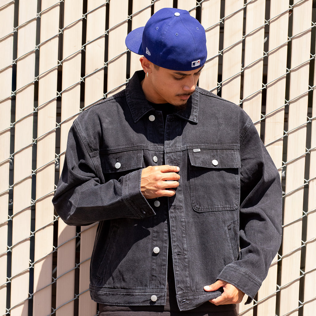 Person wearing a dark denim jacket and blue cap against a wire fence background