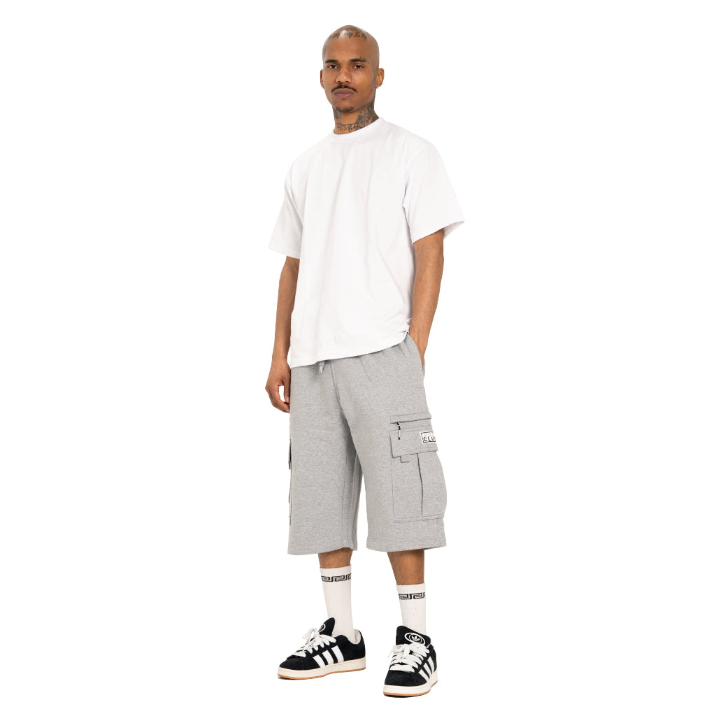 A man modeling a white oversized t-shirt, light gray cargo shorts, white socks, and black and white sneakers against a plain white background.