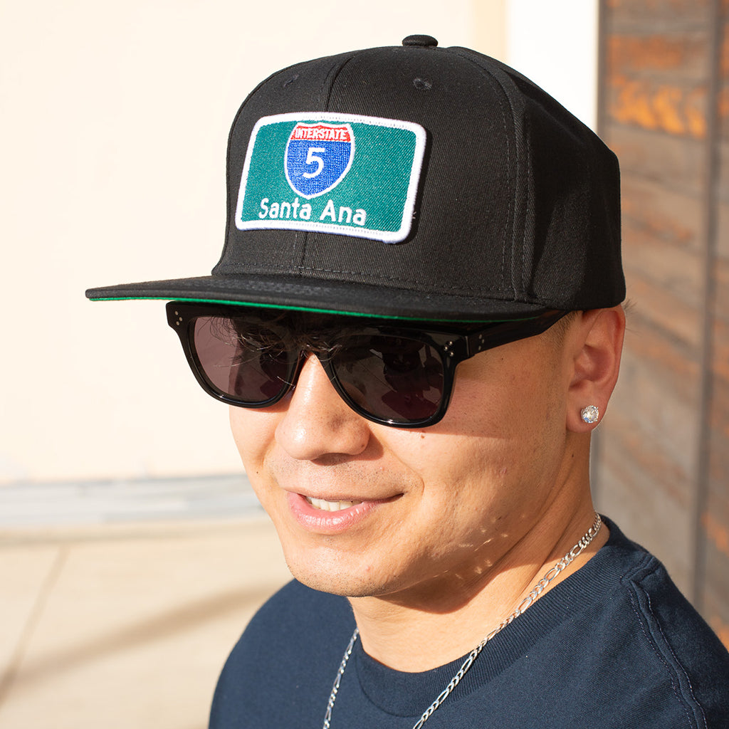 Person wearing a black cap with a 'Santa Ana' patch and sunglasses, standing against a neutral background.