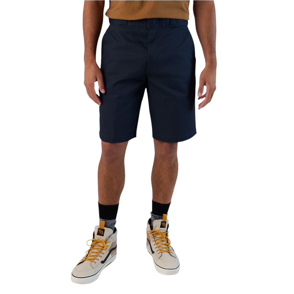 A person standing against a white background, wearing dark navy work shorts and a beige shirt.