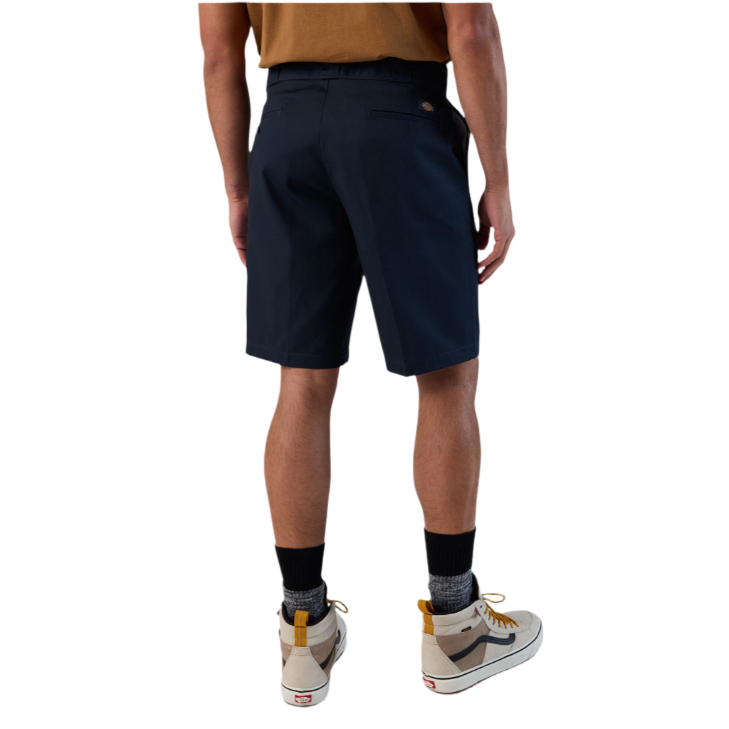 A person standing against a white background, wearing dark navy work shorts and a beige shirt.