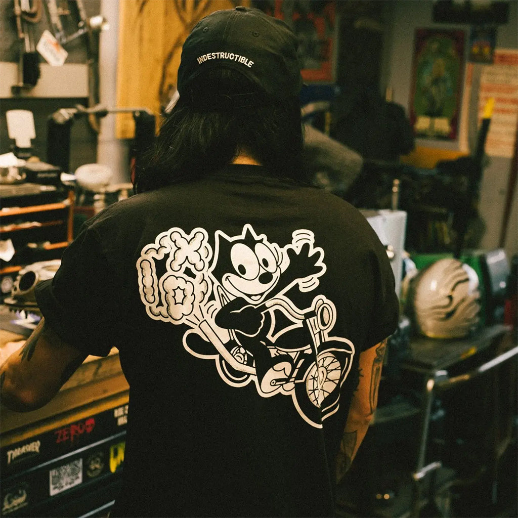 Person wearing a black t-shirt with a graphic design of a character on a bike, standing in a cluttered room.