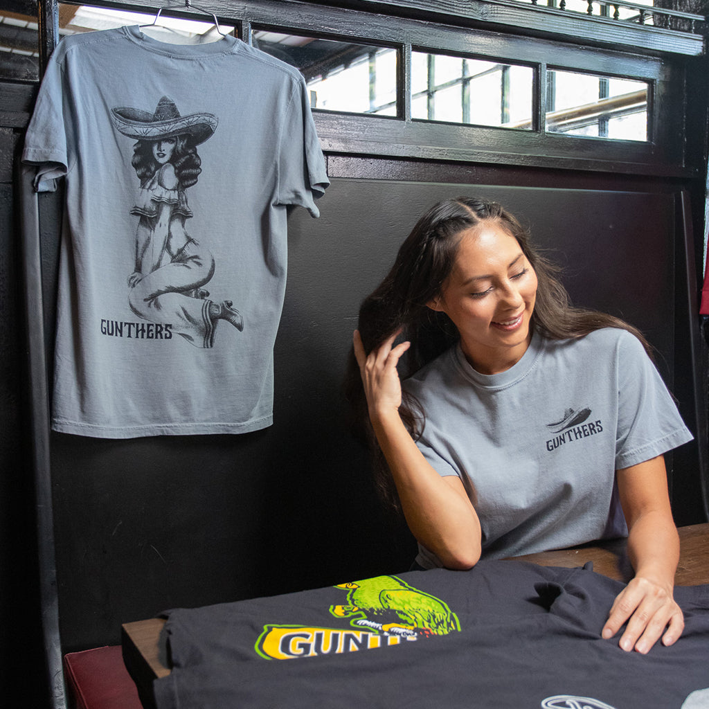 Woman wearing a gray t-shirt with a graphic design, sitting next to the Chula t-shirt with a 'GUNTHERS' logo.