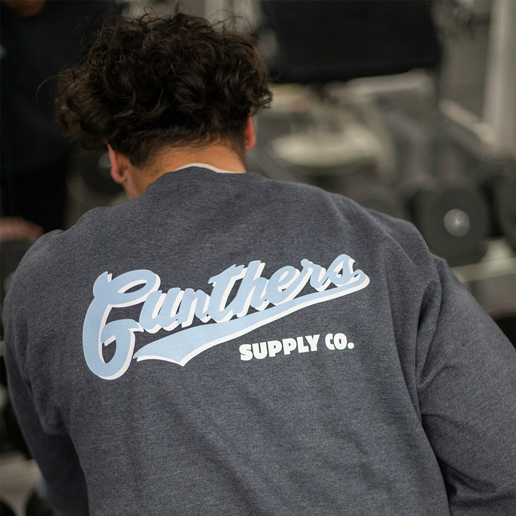 A person wearing a heather grey crewneck sweatshirt with 'Gunthers Supply Co.' text on the back.
