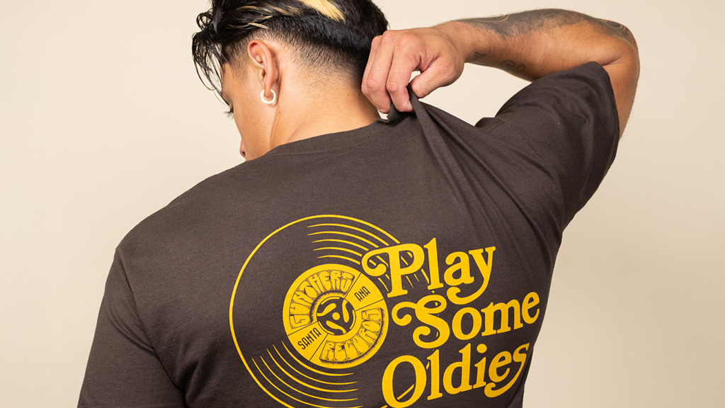 Man wearing a brown t-shirt with 'Play Some Oldies' design on the back against a beige background