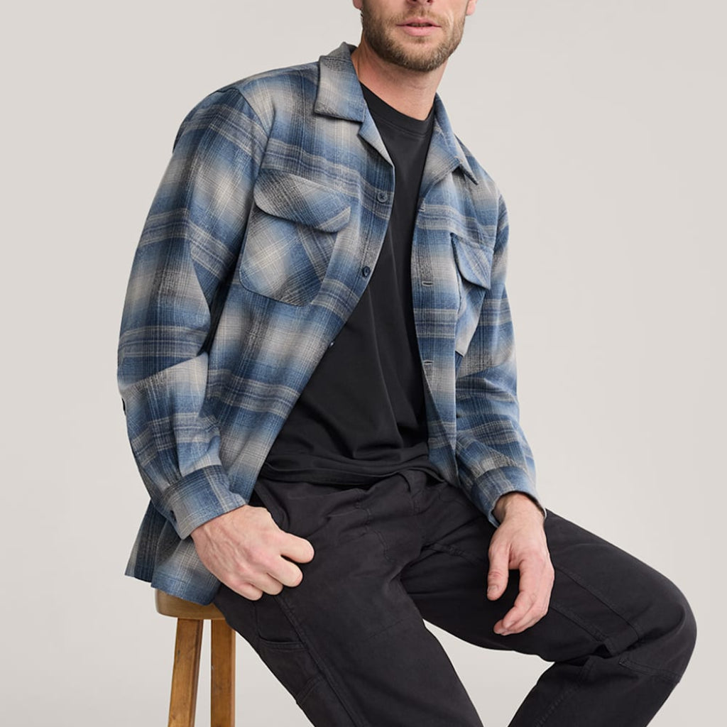 Man wearing a blue plaid shirt over a black t-shirt, sitting on a stool against a plain background.