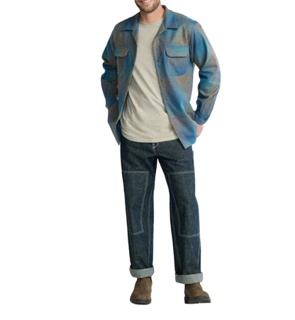 Man wearing a blue and gray shirt, beige t-shirt, and dark jeans on a light background