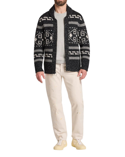 Man wearing a patterned cardigan over a plain shirt with light pants on a white background