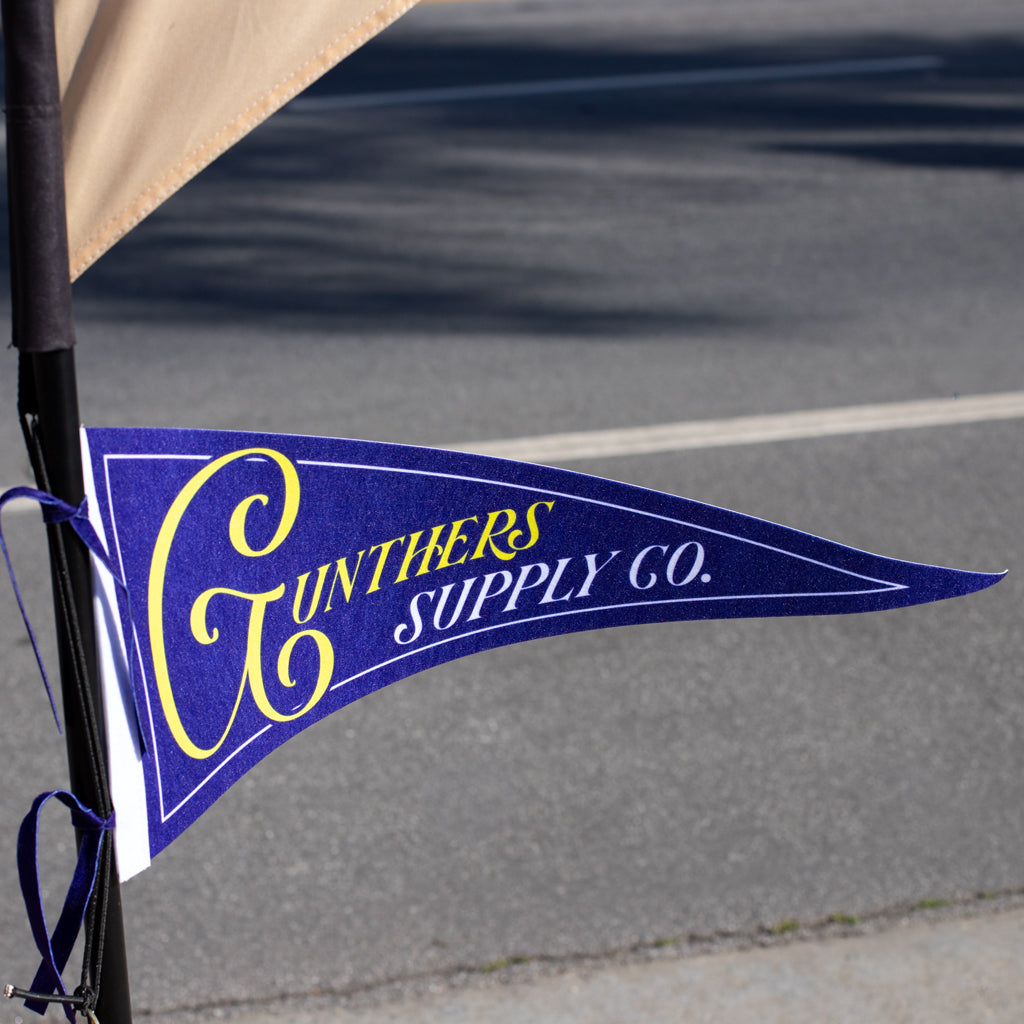 Blue and yellow pennant flag with 'Gunters Supply Co.' text on a blurred street background