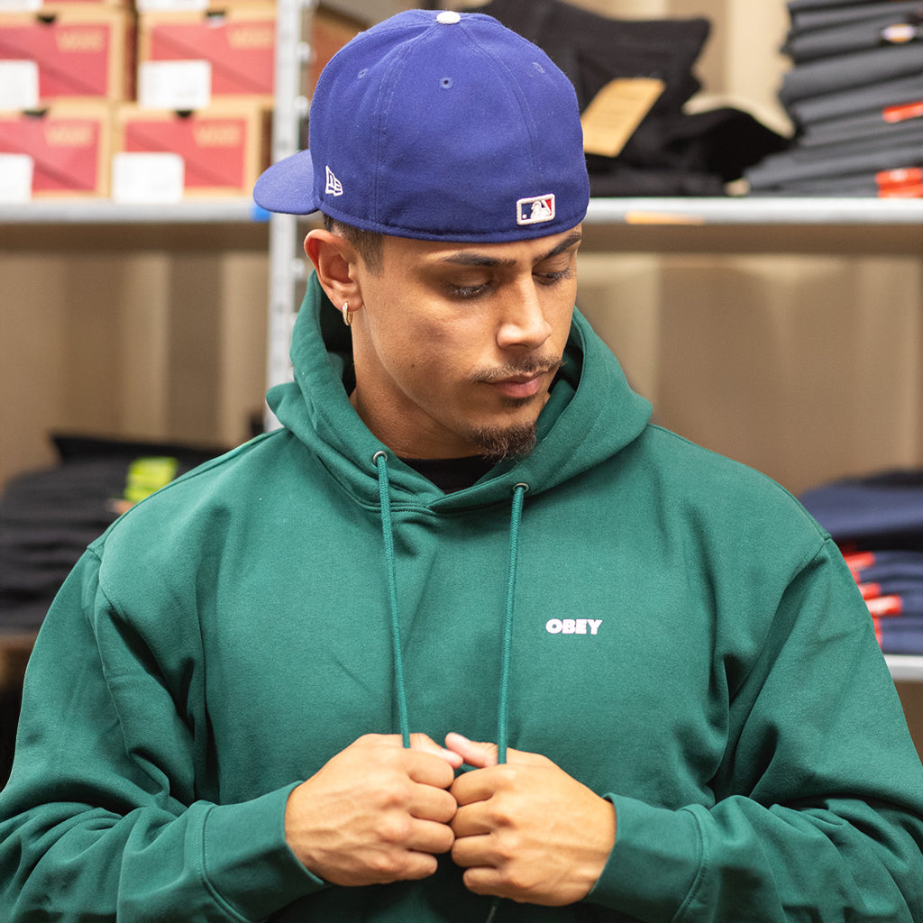 Man wearing a green hoodie with 'Obey' logo and a blue cap in an indoor setting.