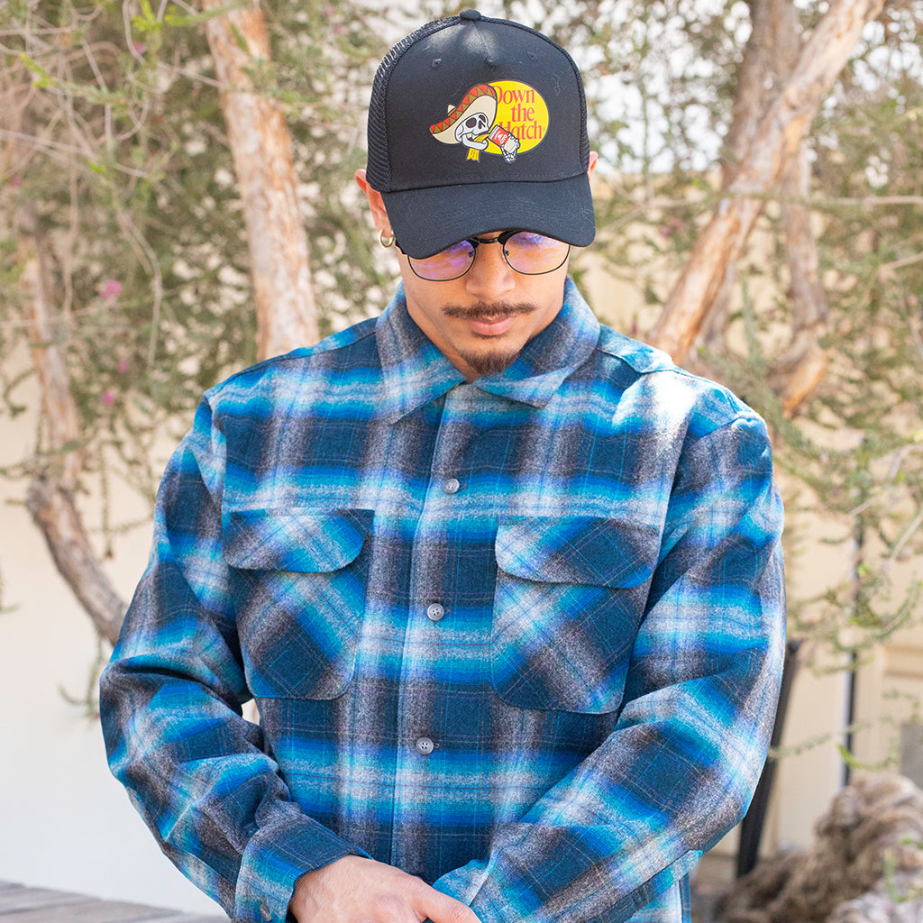 Person wearing a blue plaid shirt and a black cap with a logo outdoors.