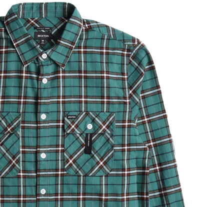 A close-up of a Brixton long-sleeve plaid shirt in green, white, and dark red, featuring a button-down collar, white buttons, and a chest pocket with a Brixton label.