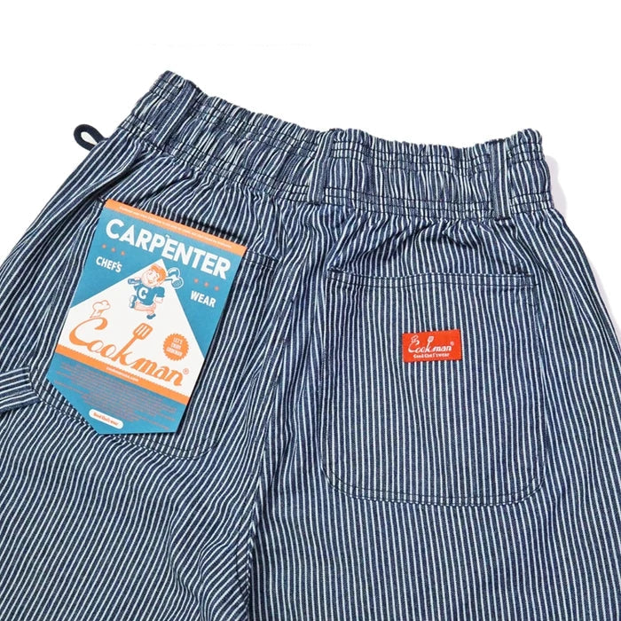 Close-up of striped Carpenter's Pants in 'Hickory Navy' by Cookman, featuring a blue and white pinstripe pattern, an elastic waistband, a side pocket with a red brand tab, and a paper hangtag.