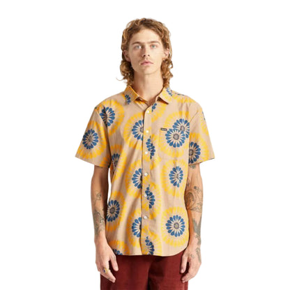 A man wearing a short-sleeve button-up shirt with a beige base and a yellow and blue sunburst pattern, paired with dark red pants, standing against a plain white background.