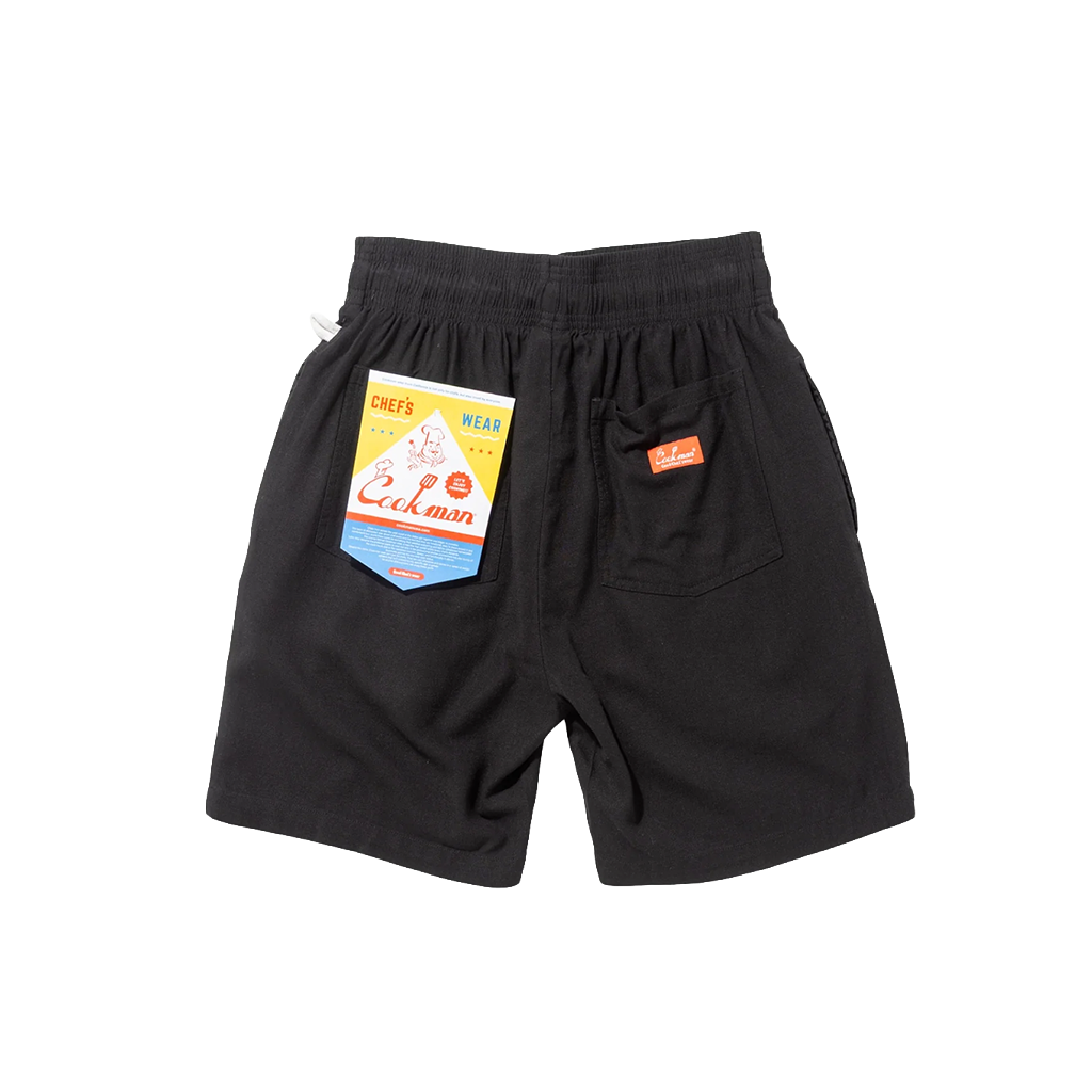 Cookman tag and red logo on the back pockets of a pair of black shorts.