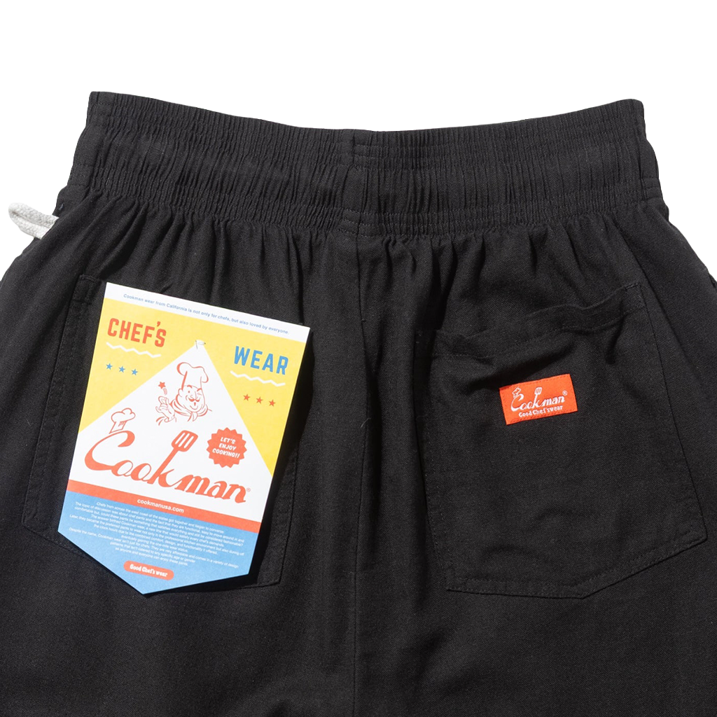 Close up Cookman tag and red logo on the back pockets of a pair of black shorts.