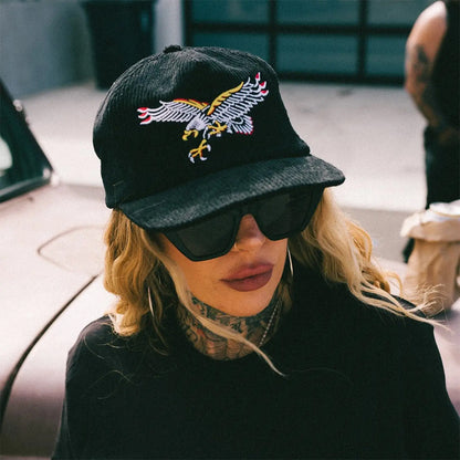 Person wearing a black cap with an eagle emblem and sunglasses, sitting in a car.