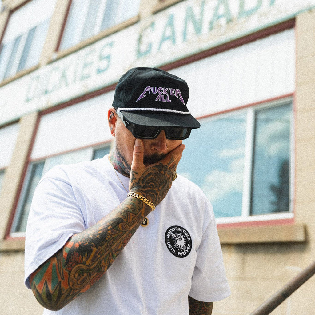 Man wearing the Fuck'em All cap and sunglasses, with tattoos on his arm, in front of a building.