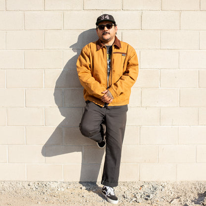 Man wearing a mustard jacket and sunglasses leaning against a beige brick wall.