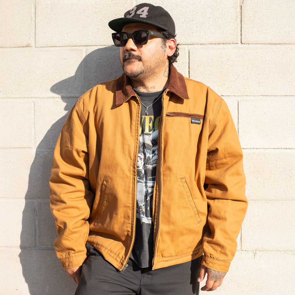 Man wearing a brown jacket and black cap against a light-colored wall