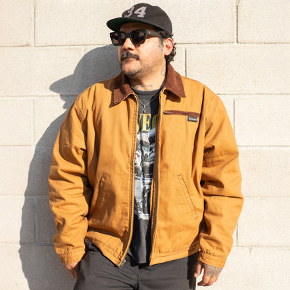 Man wearing a brown jacket and black cap against a light-colored wall