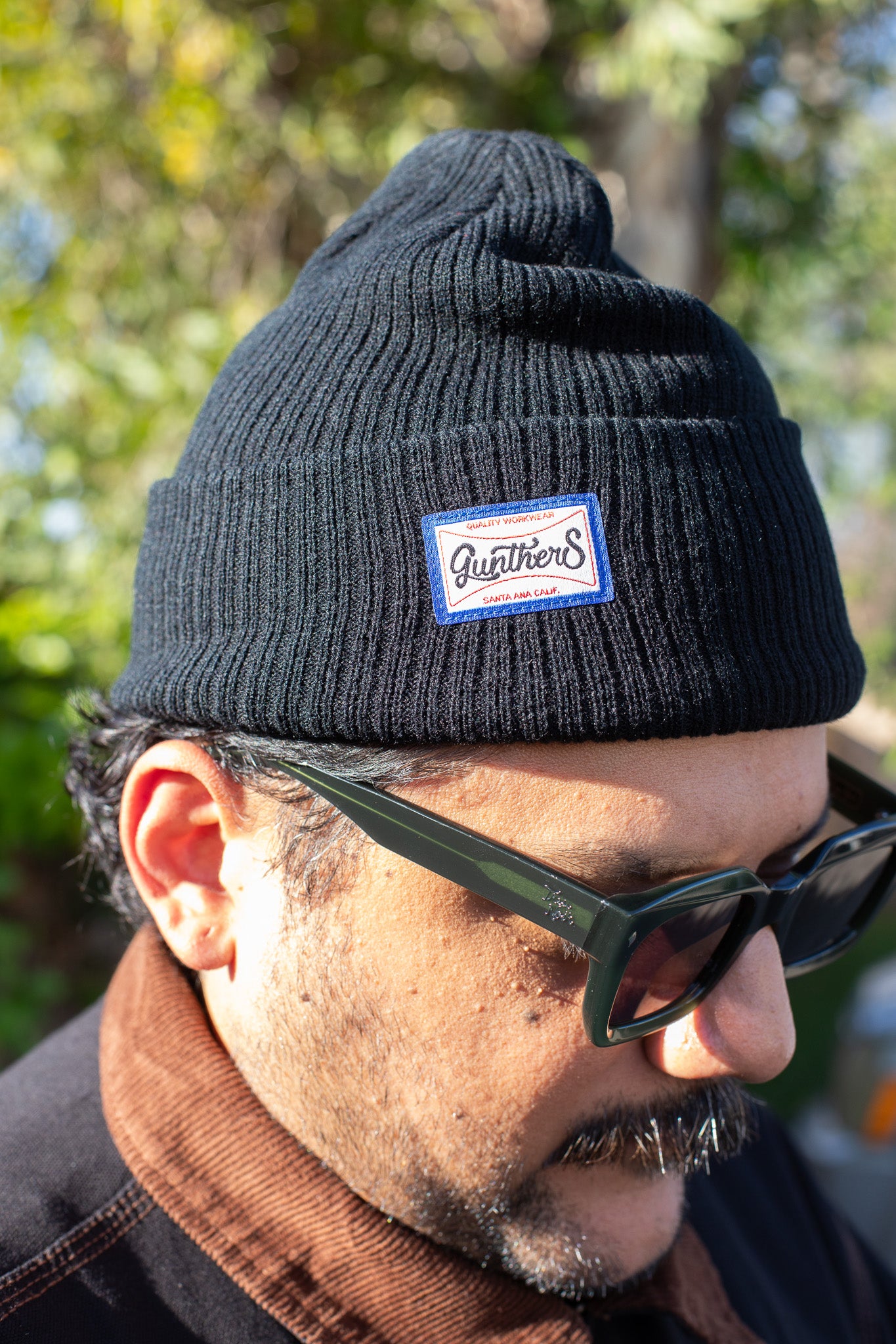 Man wearing a black beanie with a logo and sunglasses, outdoors.