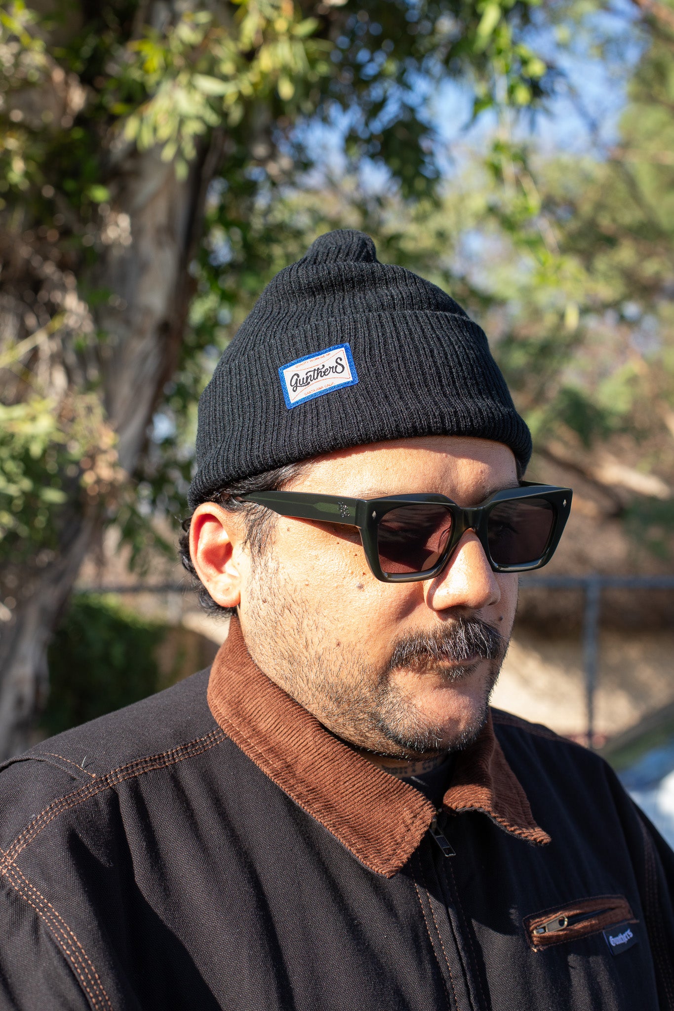 Man wearing a black beanie with a logo and sunglasses, standing outdoors with trees in the background.