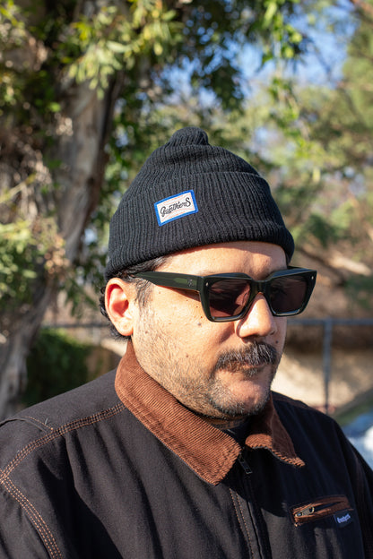 Man wearing a black beanie with a logo and sunglasses, standing outdoors with trees in the background.