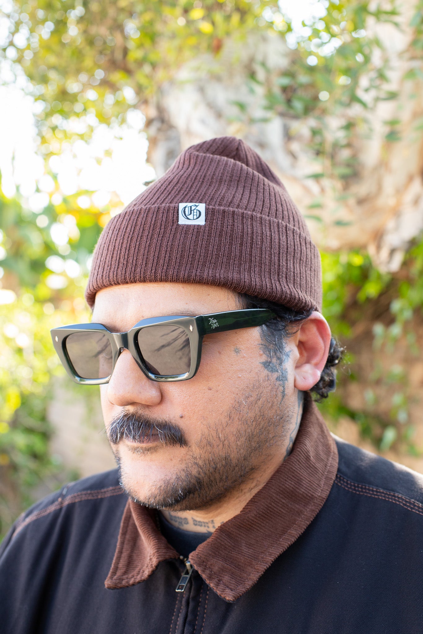 Man wearing a brown beanie with a logo and sunglasses, standing outdoors with greenery in the background.