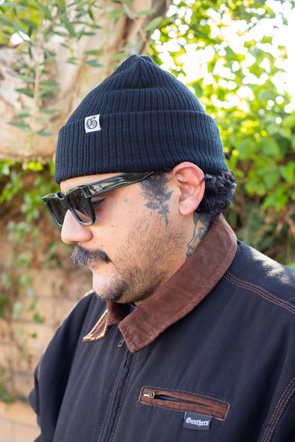 Man wearing a black beanie with a logo, sunglasses, and a dark jacket with a brown collar.
