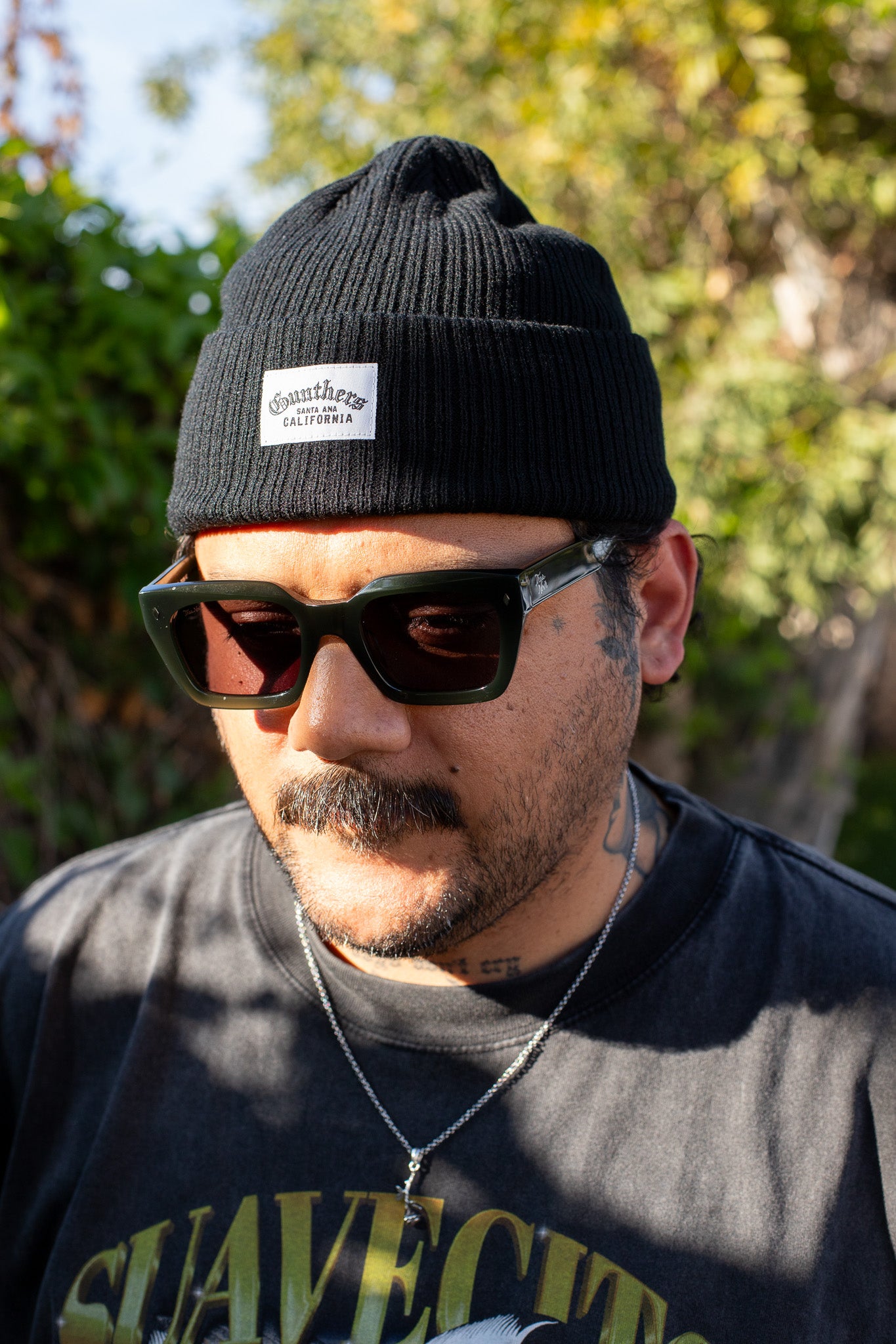Man wearing a black beanie with a logo and sunglasses, standing outdoors with greenery in the background.