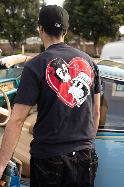Person wearing a t-shirt with cartoon characters in a heart design, standing in front of vintage cars.