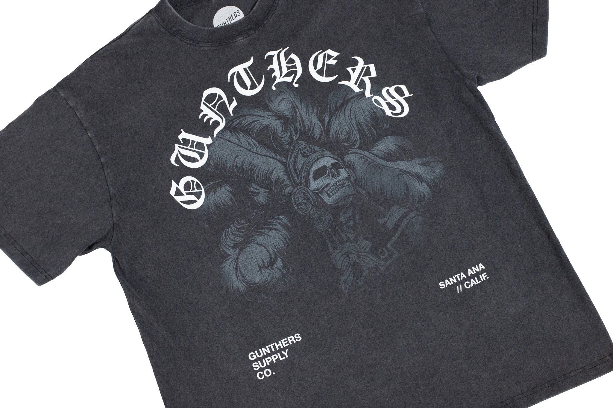 A dark gray acid-wash graphic t-shirt featuring a gothic-style white print of a skull in a feathered headdress with the word 'GUNTHERS' arched above it. Smaller white text on the lower front reads 'GUNTHERS SUPPLY CO.
