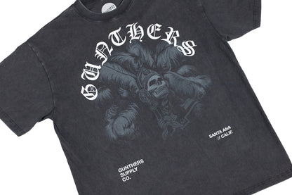 A dark gray acid-wash graphic t-shirt featuring a gothic-style white print of a skull in a feathered headdress with the word 'GUNTHERS' arched above it. Smaller white text on the lower front reads 'GUNTHERS SUPPLY CO.