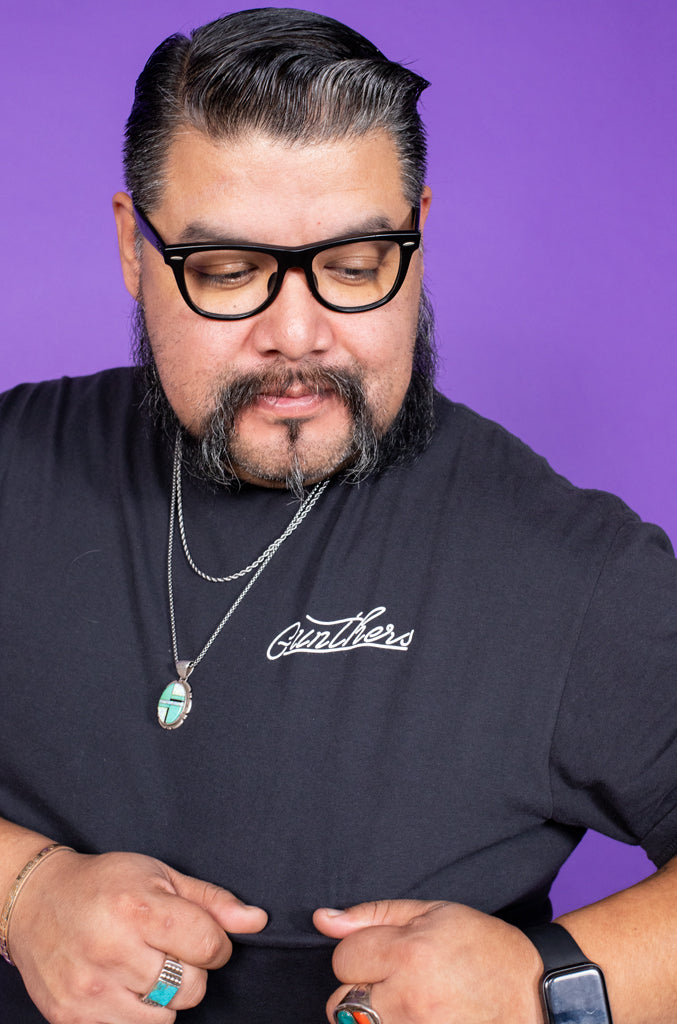 Man wearing a black t-shirt with 'Gentleman' text, glasses, and a necklace against a purple background
