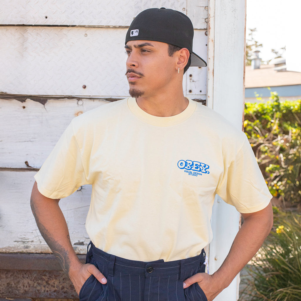 Man wearing a beige t-shirt with an 'Obey' logo, standing against a rustic wall.