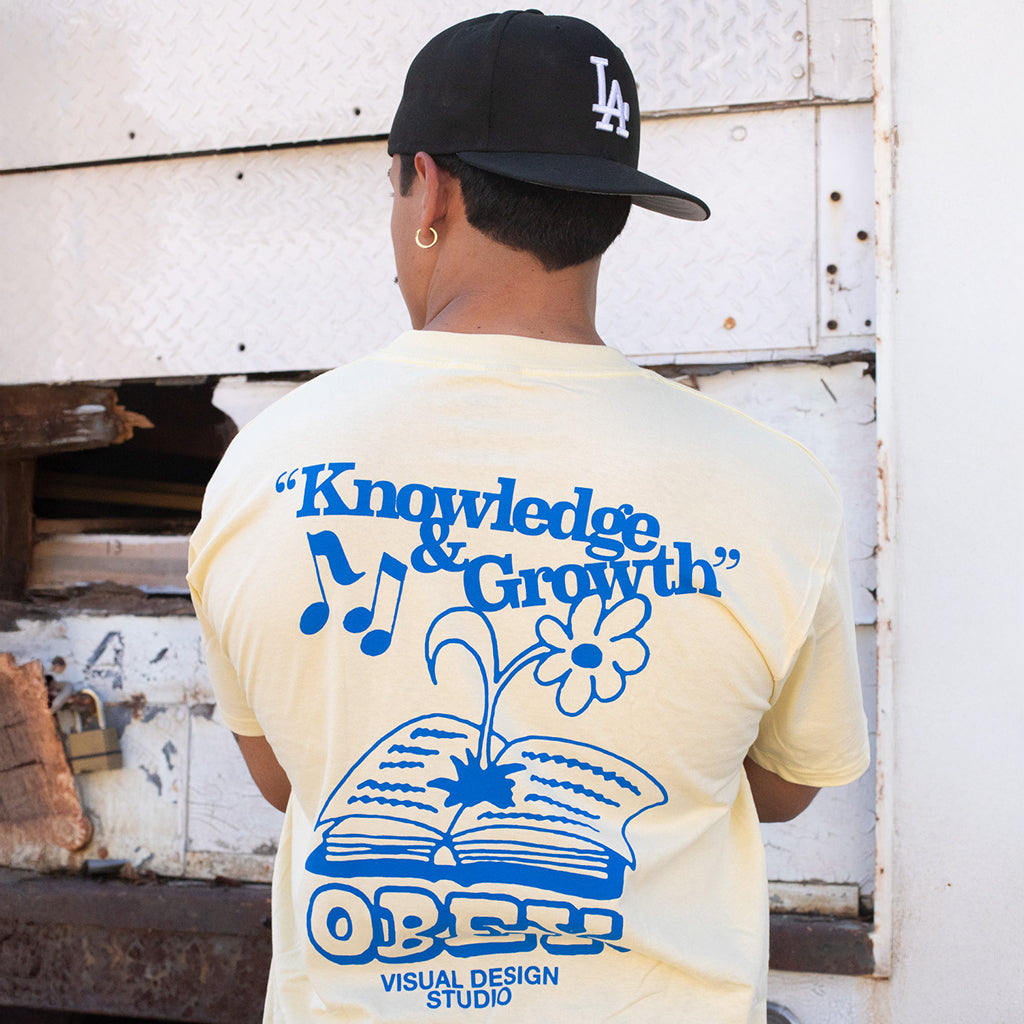 Person wearing a yellow t-shirt with blue graphics and text, standing in front of a white wall.